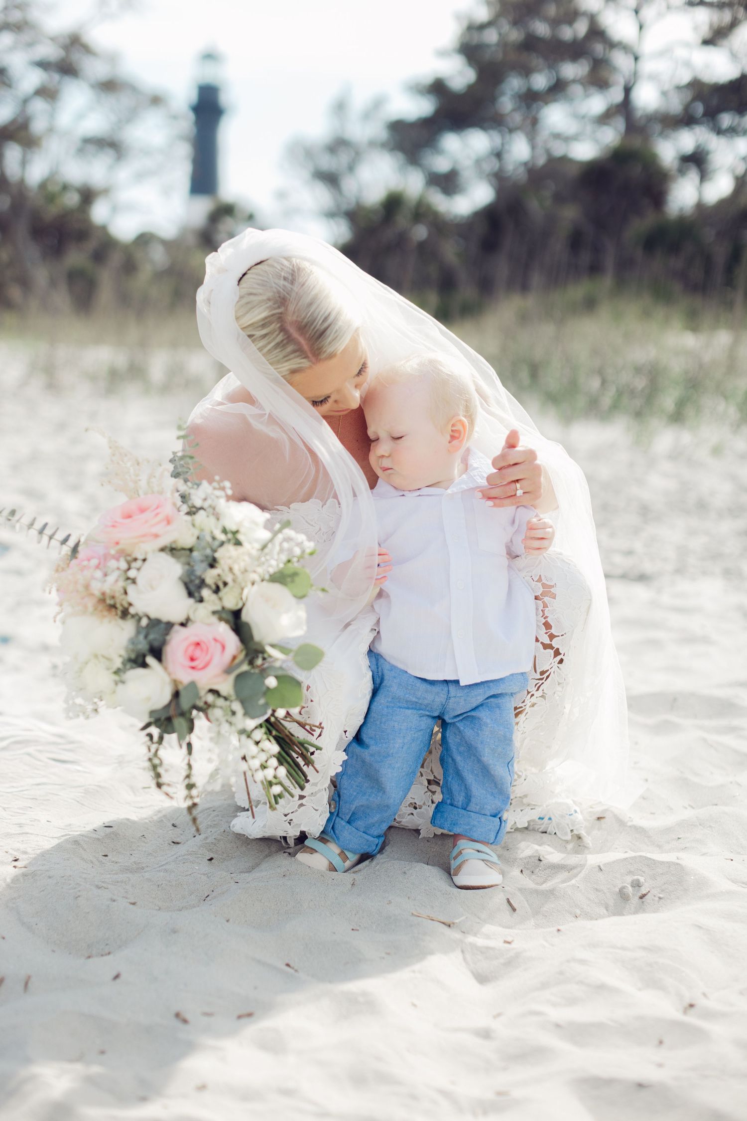 Bride in white veil kneeling on sandy beach holding bouquet, embracing toddler in blue jeans near lighthouse.