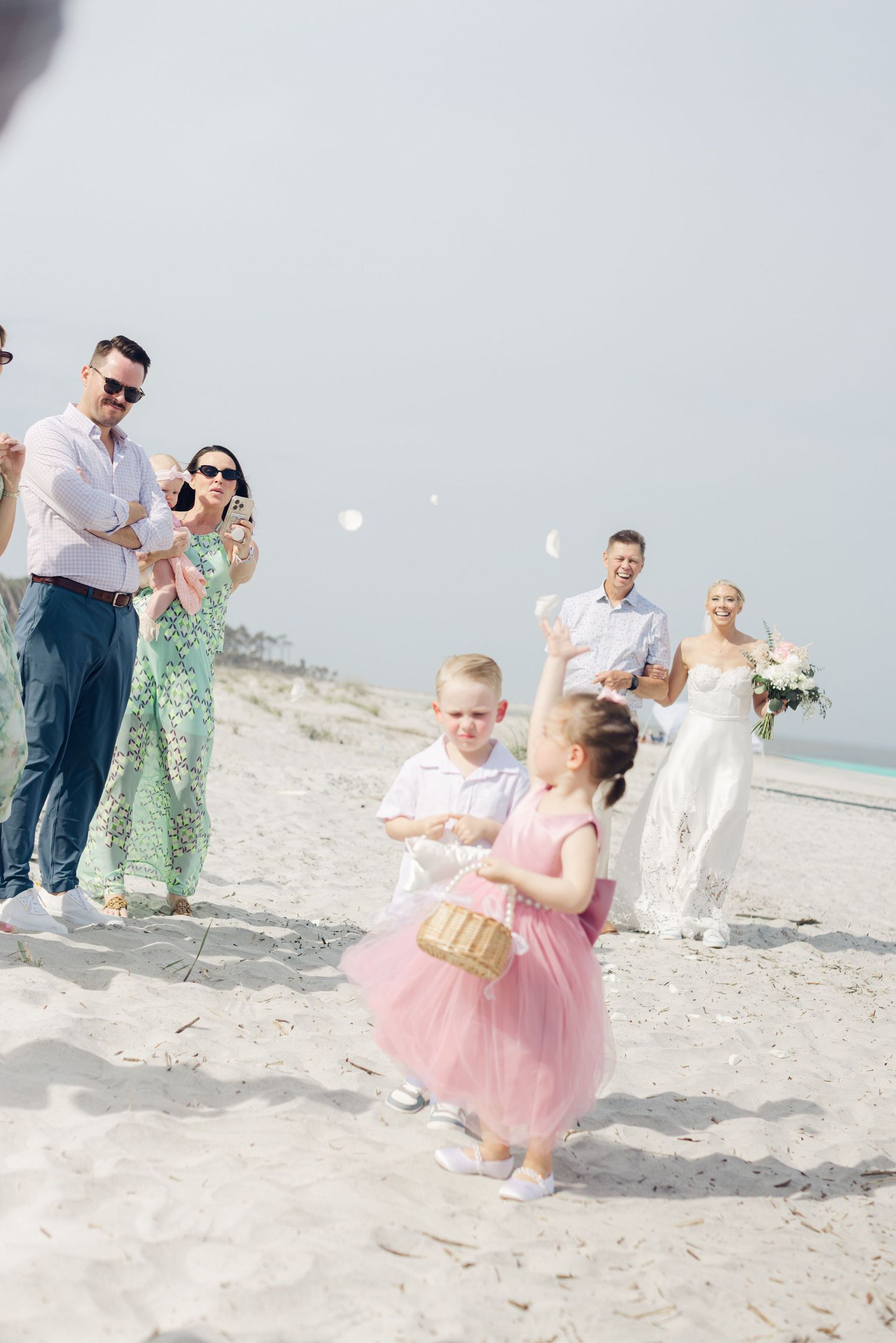 Children toss flower petals at a beach wedding while guests watch the bride and groom approach on sandy shore.