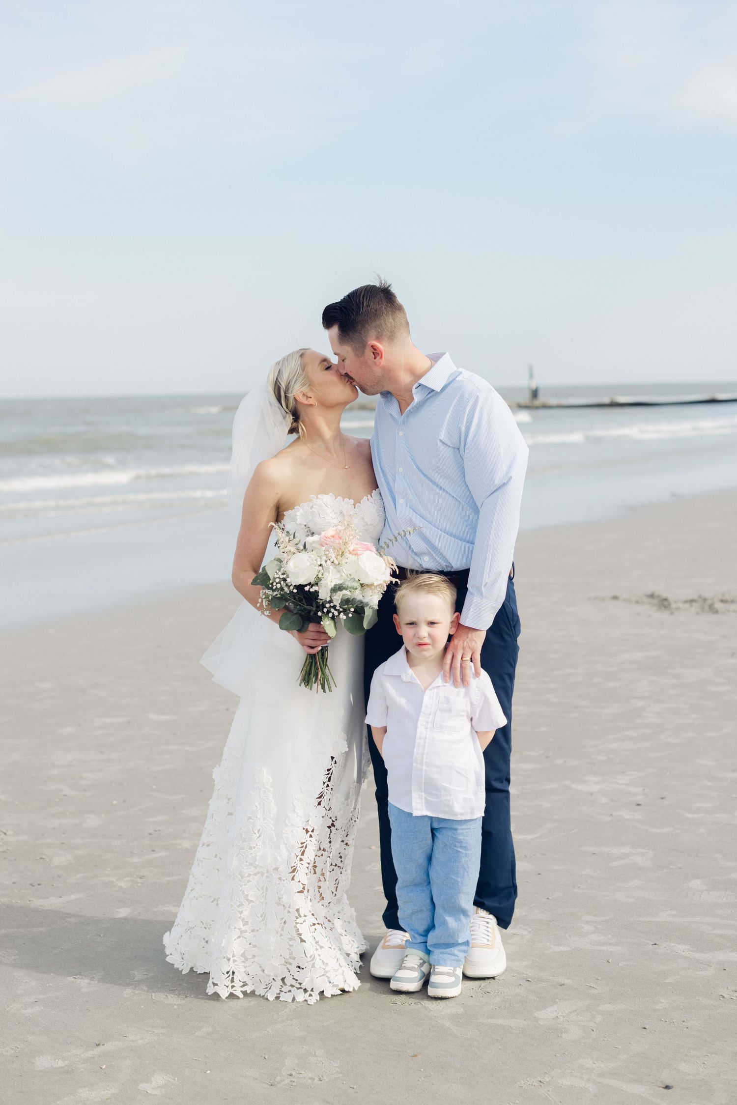 Bride and groom kissing on beach with young child, bride in lace dress holding bouquet, ocean backdrop.