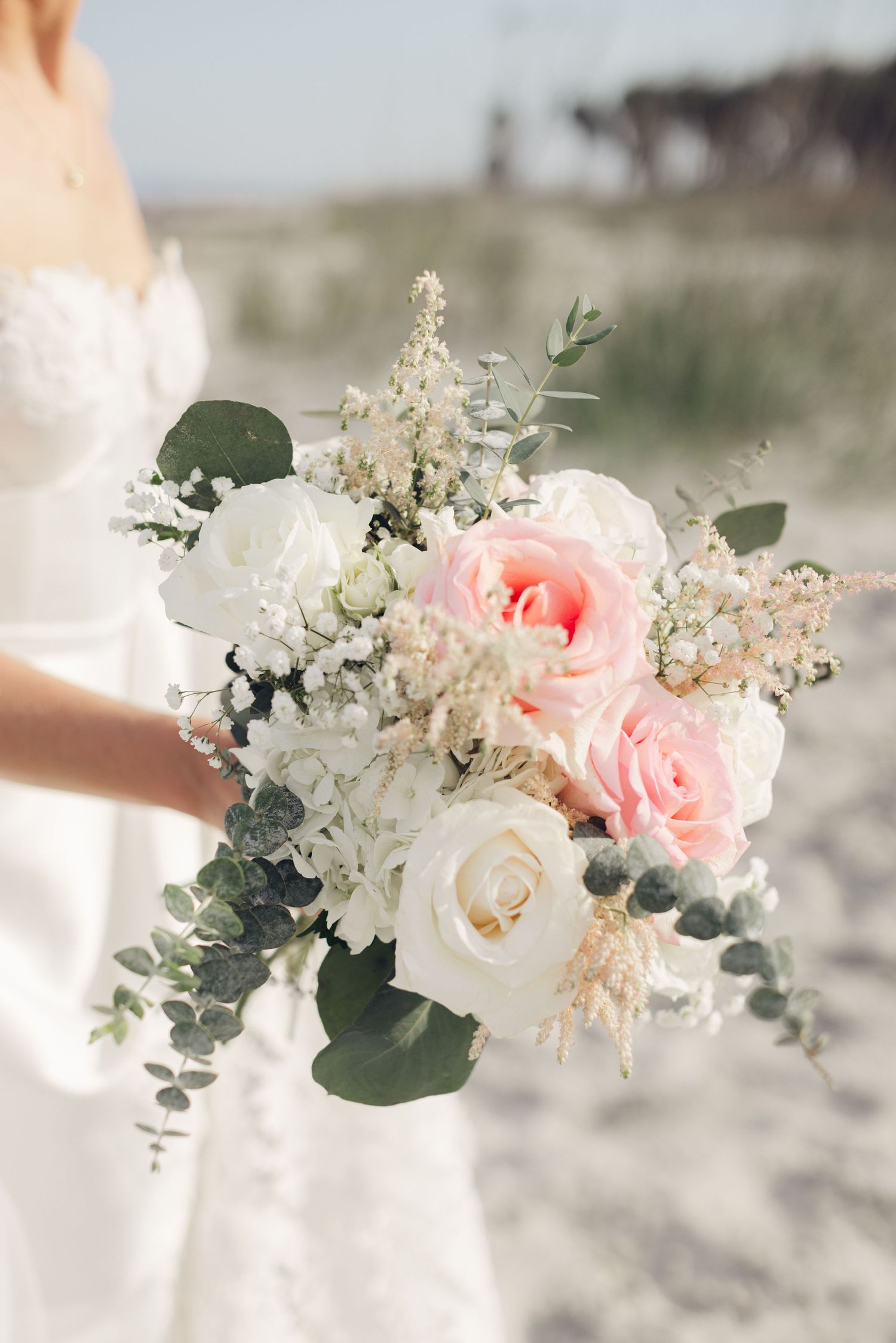 Bride holding a romantic wedding bouquet with pink and white roses, eucalyptus, and delicate blooms on a beach.