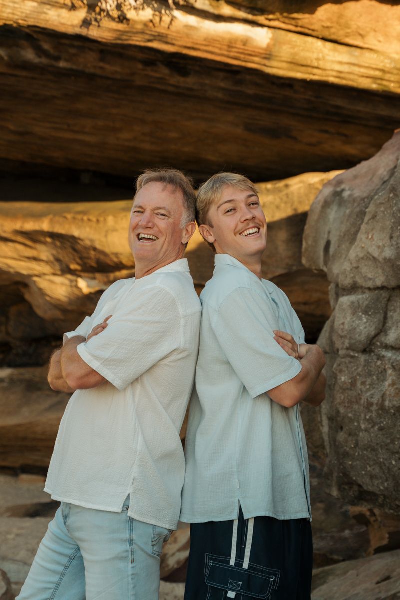 Two smiling men, son and dad in white shirts stand back-to-back with arms crossed near rocky cliffs Bronte Beach Sydney Photoshoot