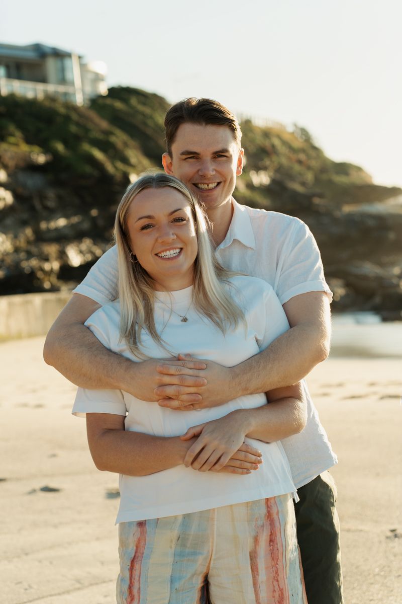 A smiling couple hugs on a beach at sunset, man in white shirt hugging woman from behind at Bronte Beach Sydney photoshoot