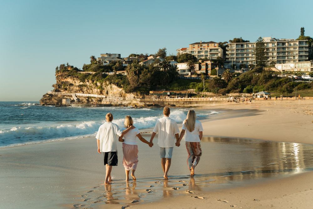 Family of four walking hand-in-hand along a sunny beach with cliffside buildings in the background Bronte Beach family photoshoot