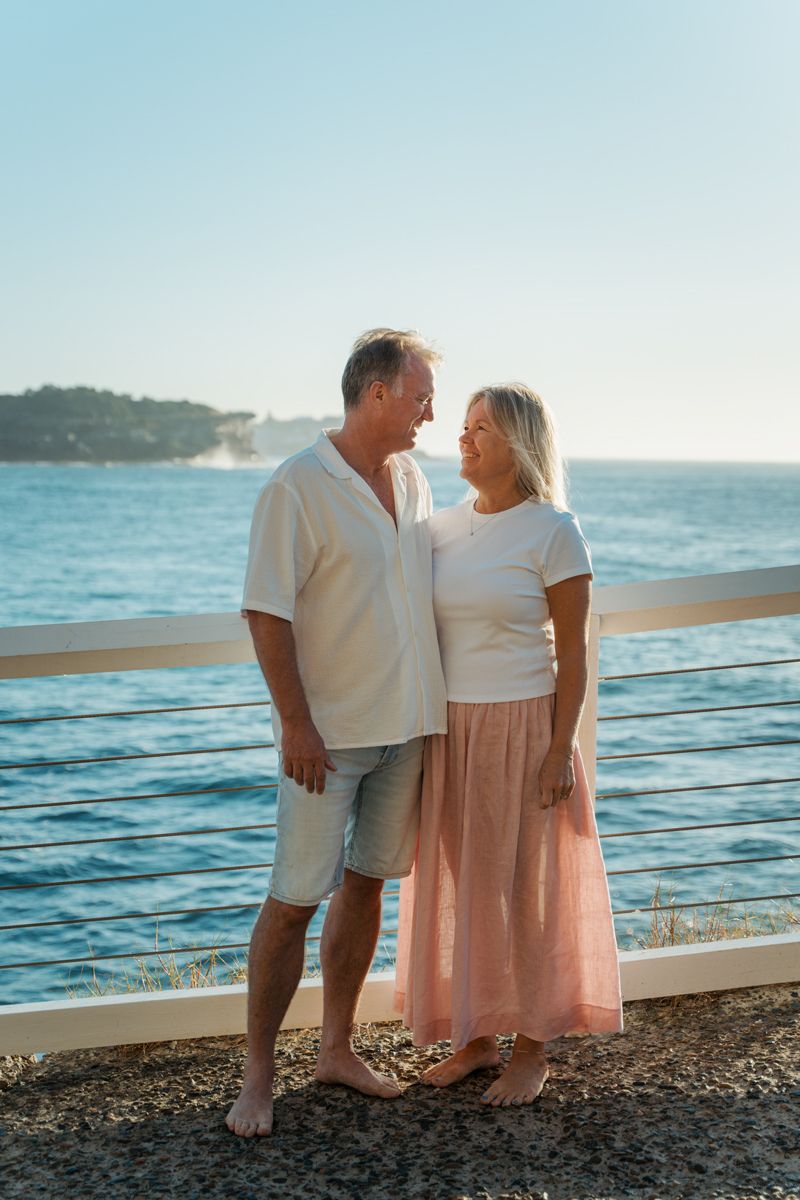 middle aged couple standing barefoot by the sea, smiling at each other in white tops and casual attire on a sunny day photoshoot Bronte