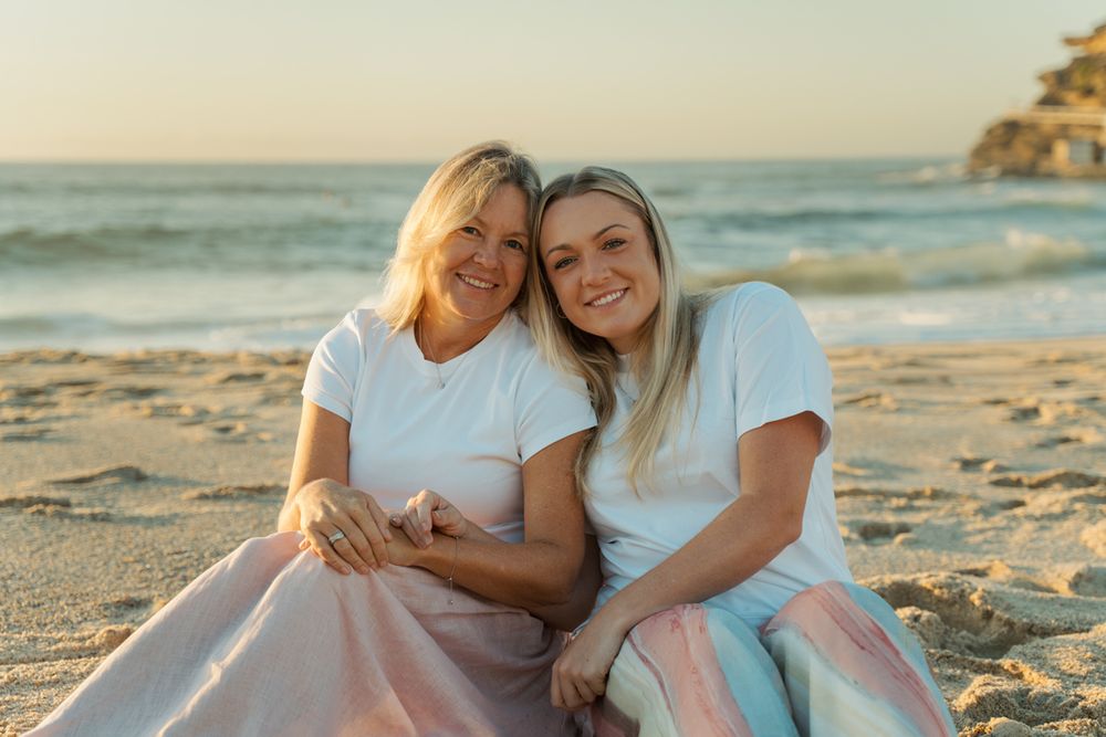 Two blonde women, mom and daughter in white tops smile together on a sandy beach at sunrise, sitting close family photoshoot at Bronte Beach
