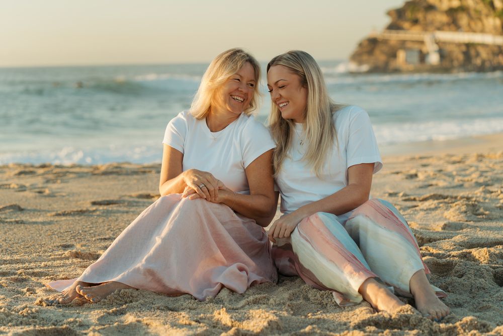 Two blonde women mom and daughter smile and sit together on a sandy beach at golden hour family photoshoot at Bronte Beach
