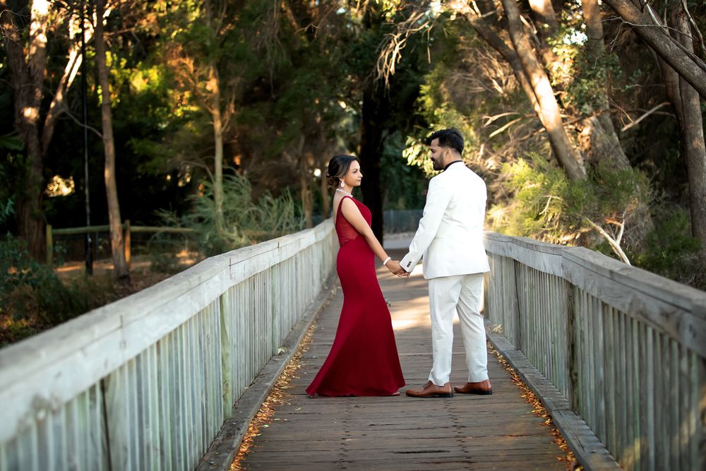 Couple holding hands on wooden bridge, woman in red gown, man in white suit, surrounded by lush green trees.