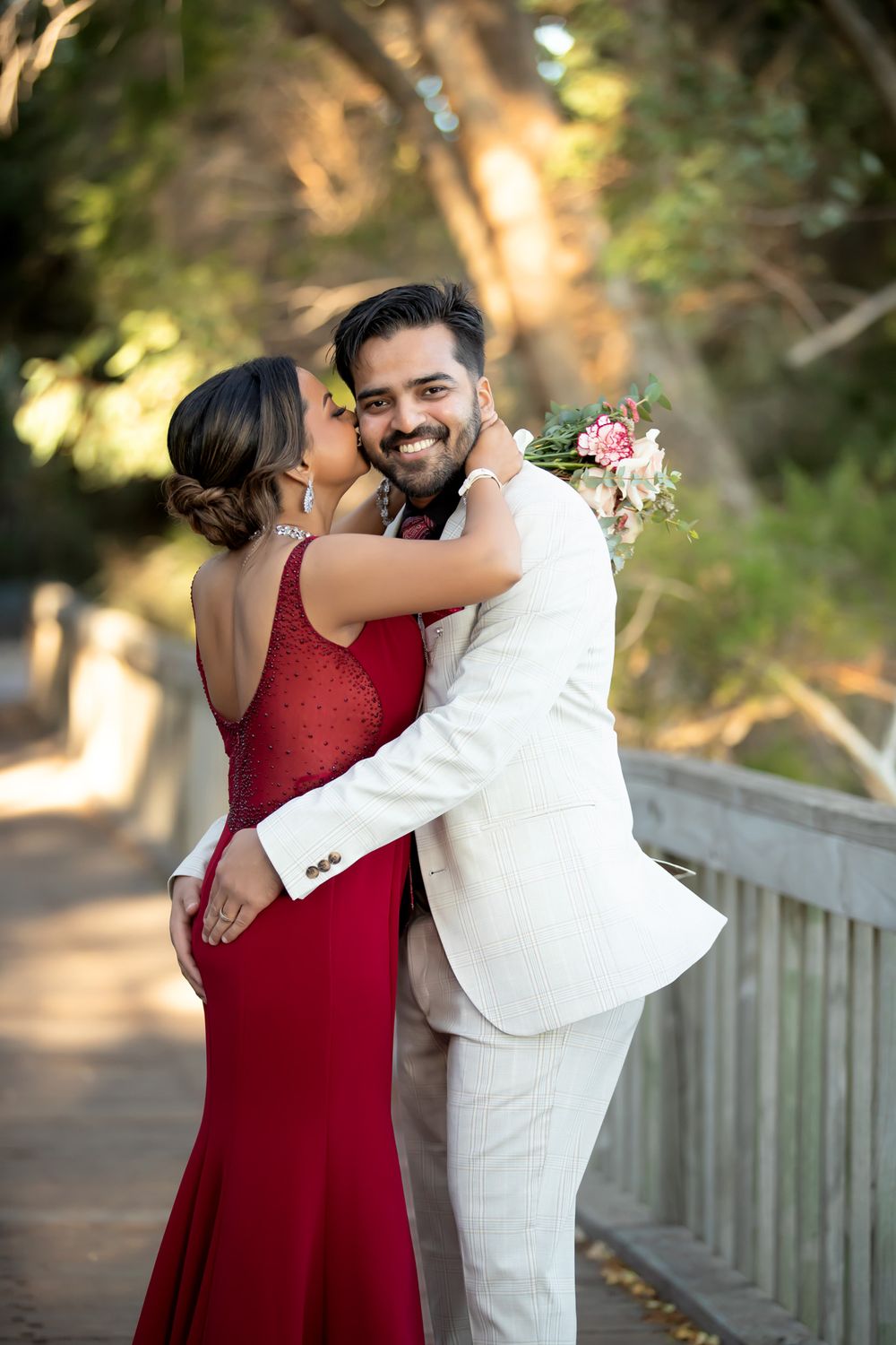 Couple embracing on a wooden bridge, woman in red gown holding bouquet, man in white suit smiling at camera.