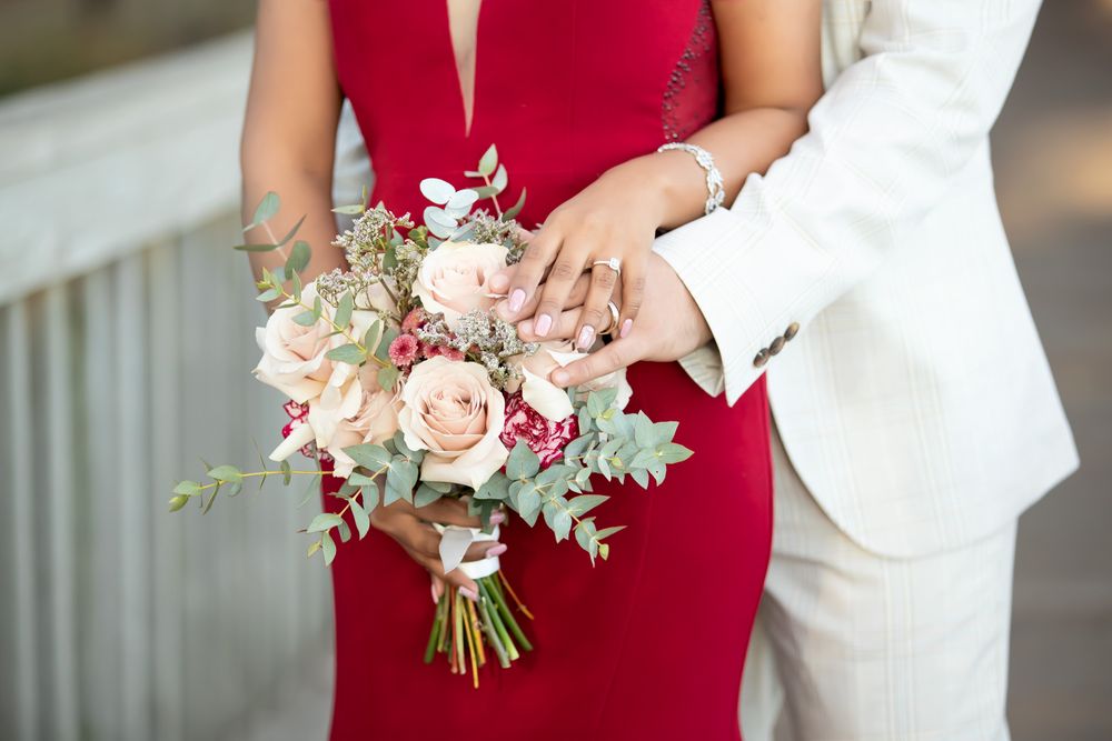 Couple in red dress and white suit holding a blush pink rose wedding bouquet with greenery.