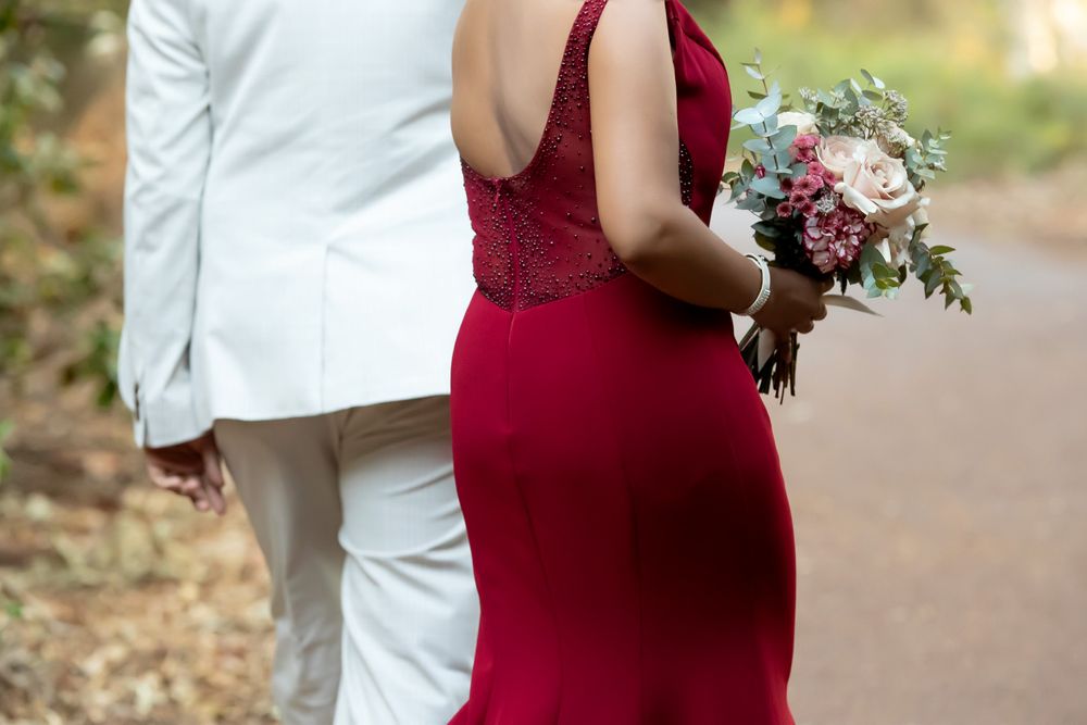 Bride in red gown holding floral bouquet stands beside groom in white suit on outdoor path.