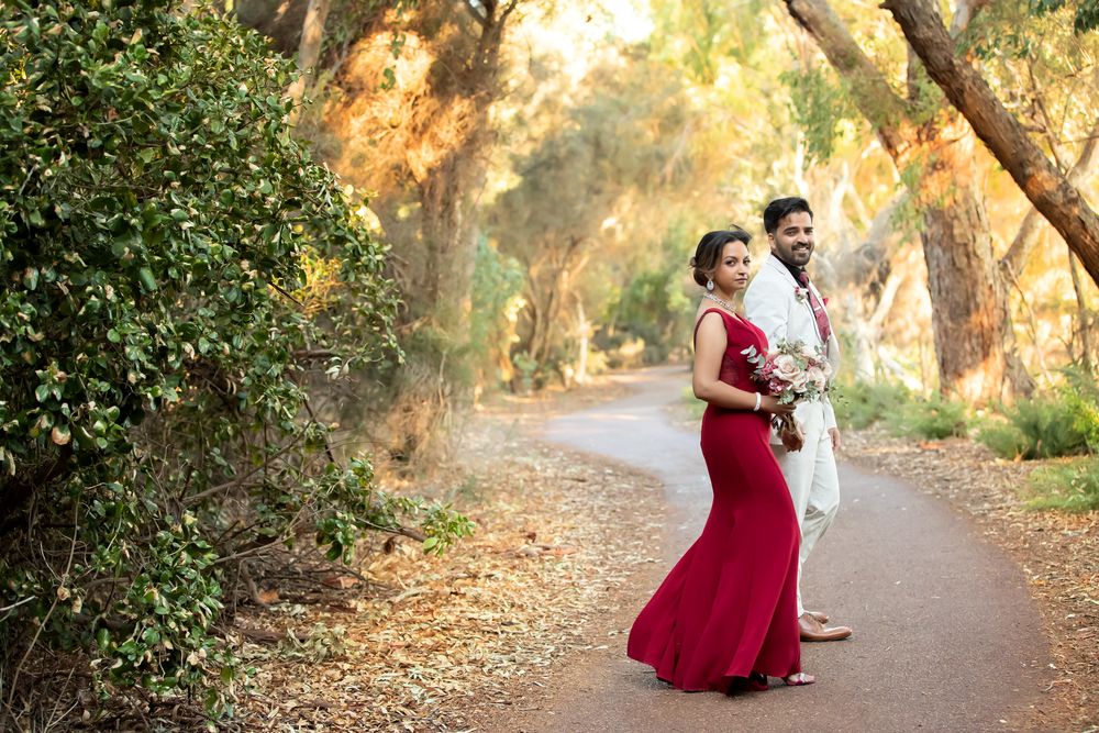 Couple in formal attire poses on a tree-lined path, bride in red gown holding bouquet, groom in white suit.
