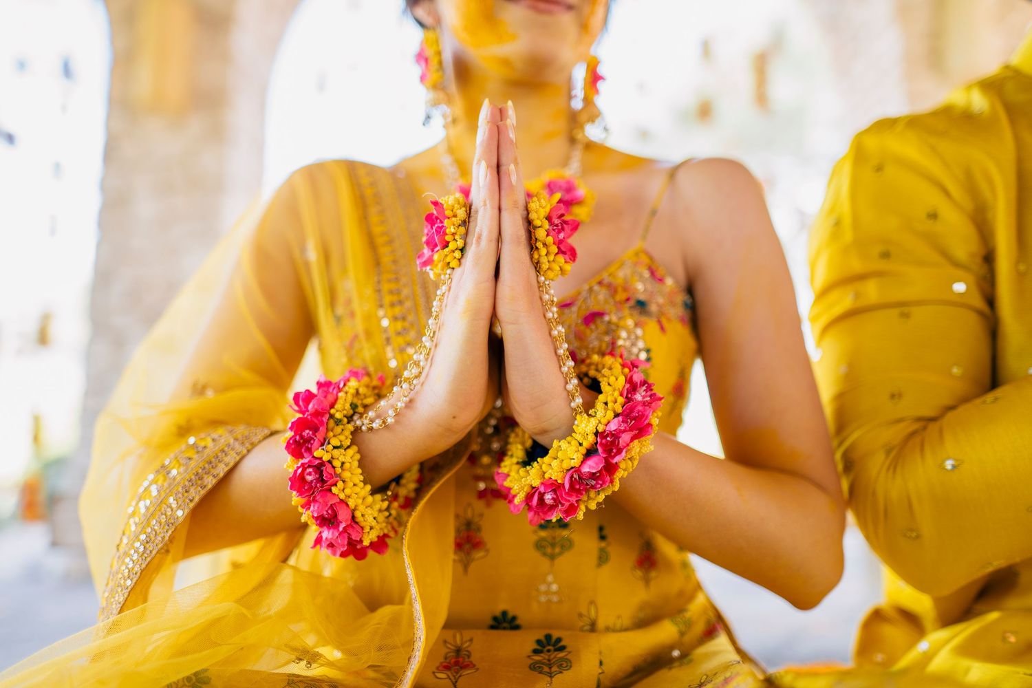 Woman in golden yellow sari performing namaste gesture with floral garlands during traditional Indian ceremony.
