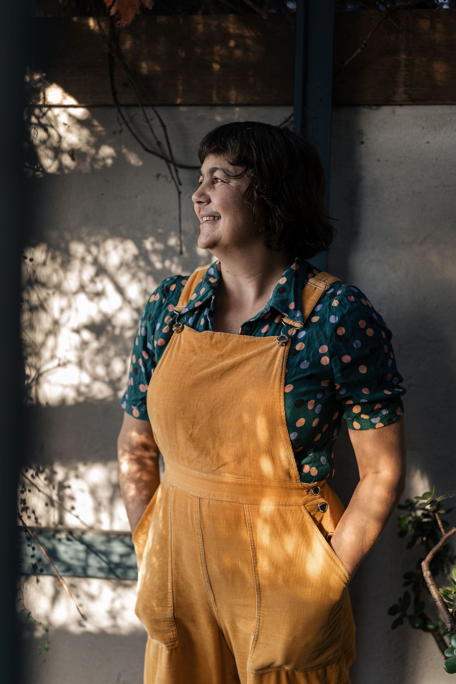 A smiling woman in yellow overalls and polka dot shirt stands in dappled sunlight outdoors.

Mara Ripani