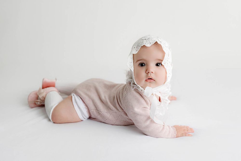 A cute baby in a pink romper and white lace bonnet lies on tummy on a white background.