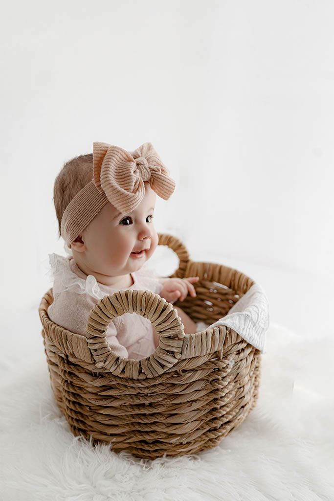 Smiling baby girl in a wicker basket wearing a pink bow headband and white lace outfit on a white background.