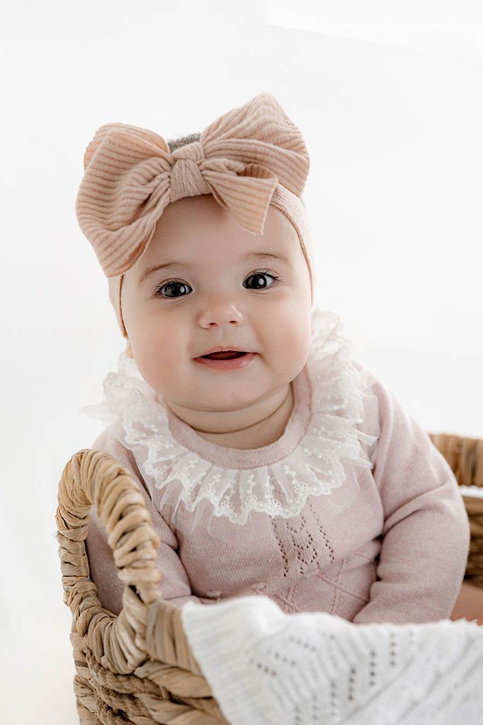 Adorable baby girl in pink knit outfit with lace collar and bow headband sitting in wicker basket.