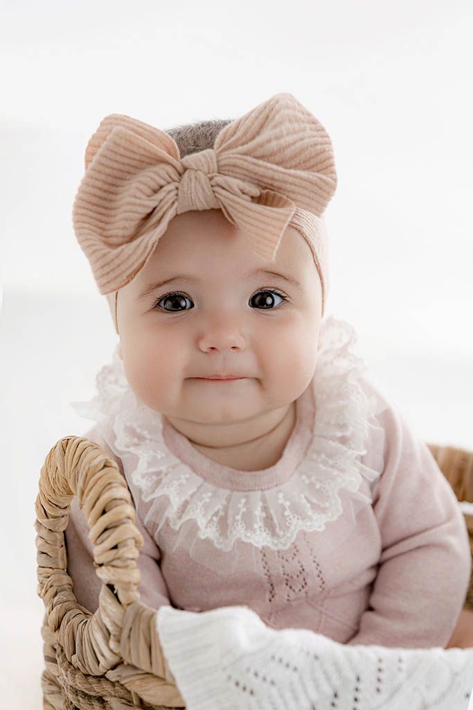 Adorable baby girl in pink knit outfit and beige bow headband sitting in a wicker basket against white background.