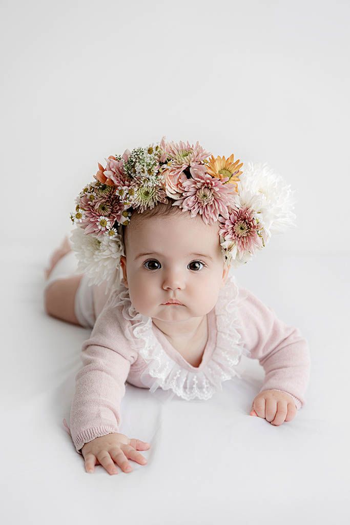 Baby girl in pink cardigan wearing a floral crown lies on white background.