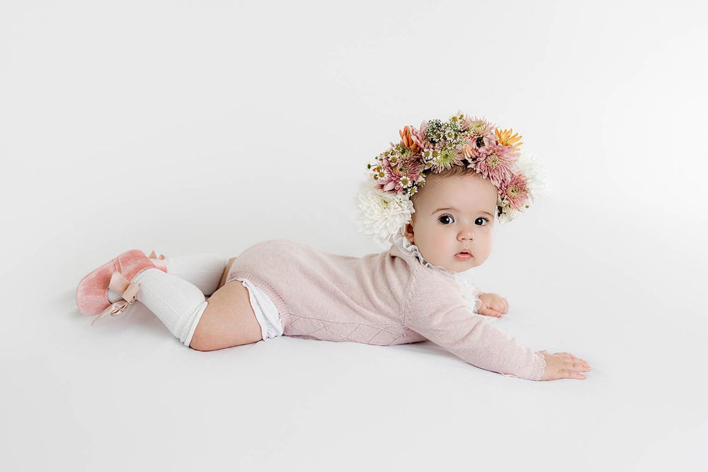 Baby girl in pink onesie and floral crown lying on white background.