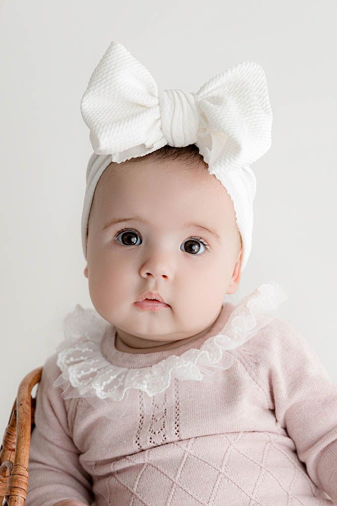 Adorable baby girl in pink knit outfit with white bow headband sits in wicker chair.