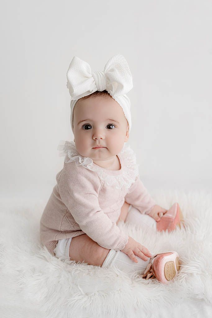 Adorable baby girl in pink sweater and white bow headband sitting on fluffy white rug with pink shoes.