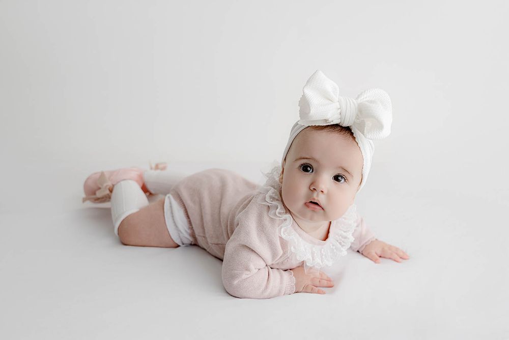 Adorable baby girl in pink onesie and white bow headband doing tummy time on white background.