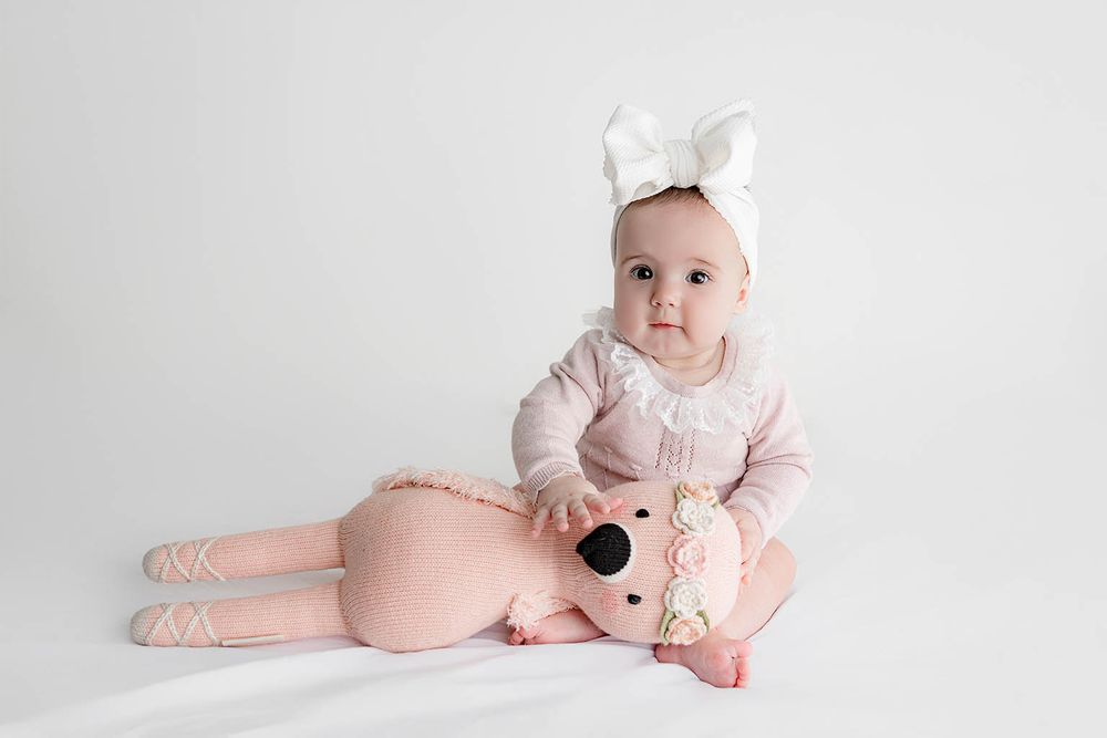Adorable baby girl in pink knit outfit and white bow headband sitting with a plush pink bear toy.