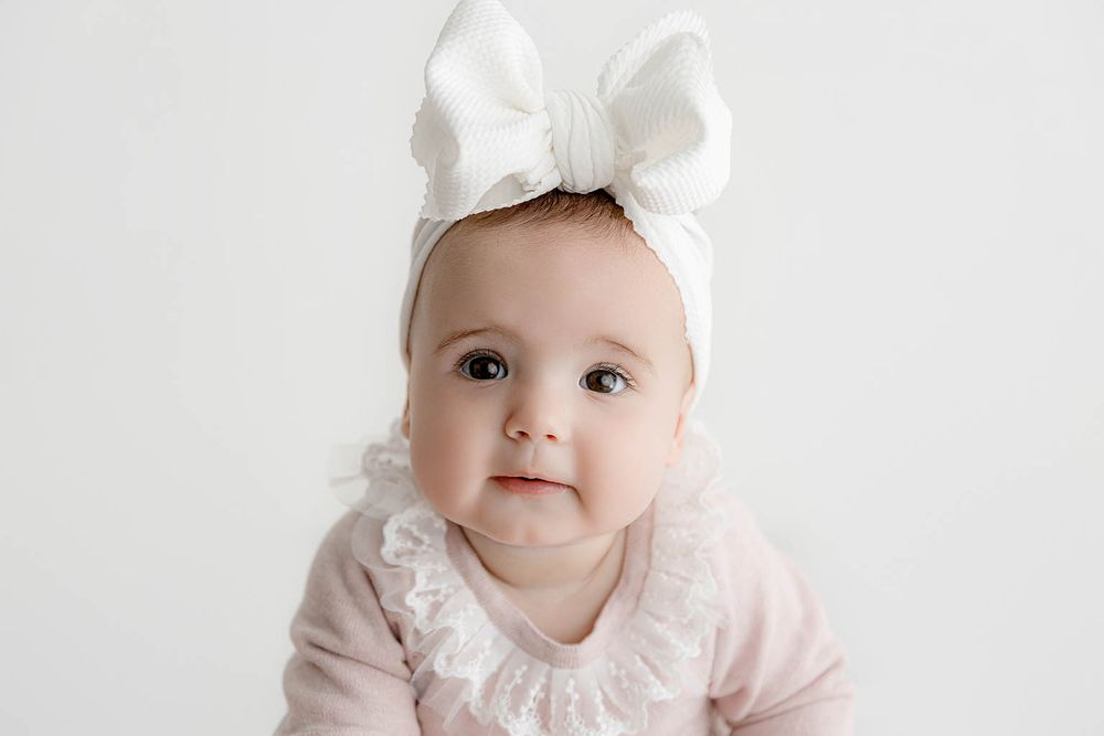 Cute baby girl wearing a white bow headband and pink ruffled outfit against a white background.