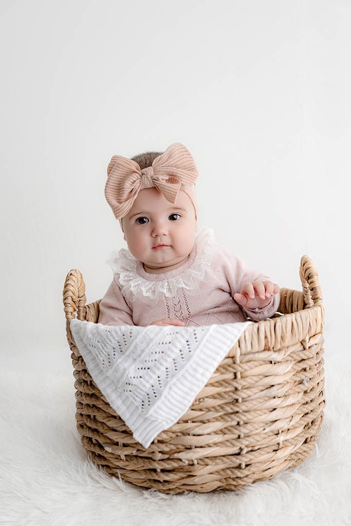 Cute baby girl in pink outfit with bow headband sits inside large wicker basket.