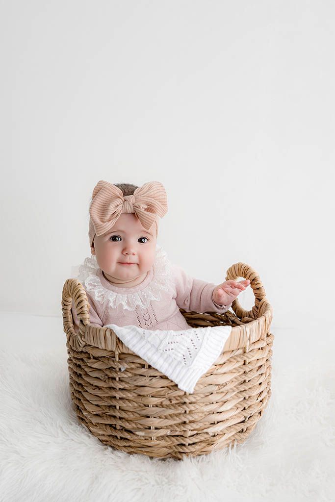 Sweet baby girl in pink knit outfit sitting inside a woven wicker basket with a white blanket.