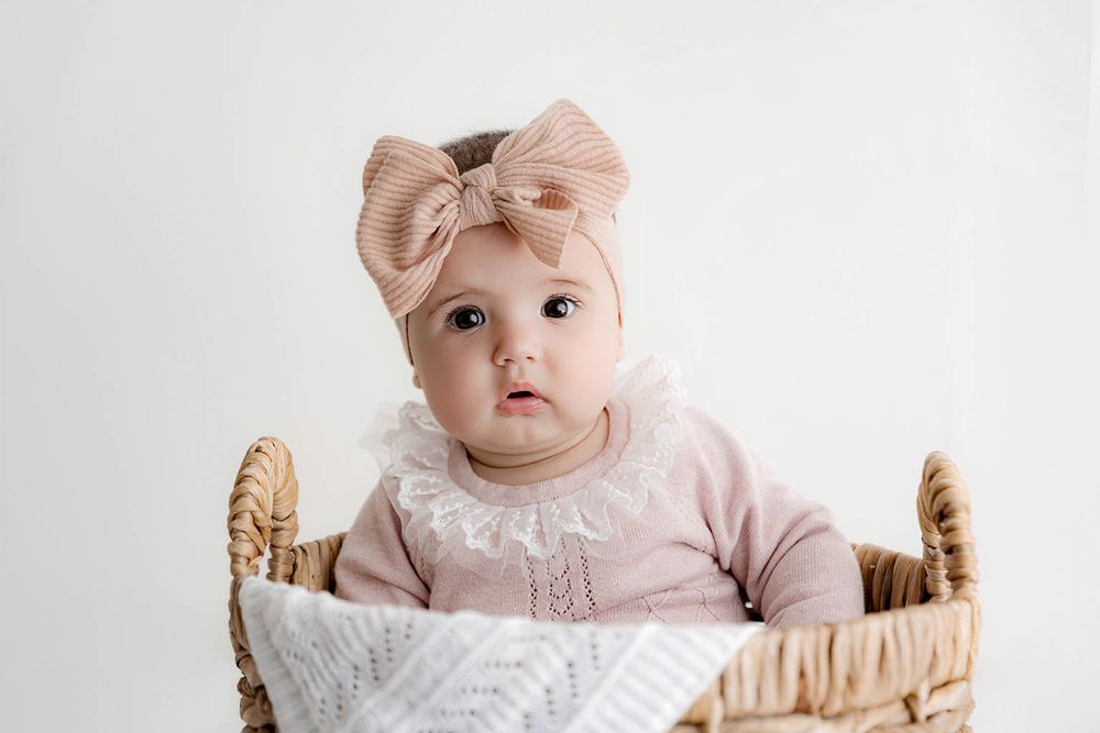 Sweet baby girl in a pink knit sweater and blush bow headband sitting in a wicker basket.