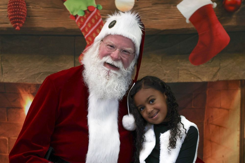 Santa Claus posing with a young girl in front of a festive fireplace with Christmas stockings.