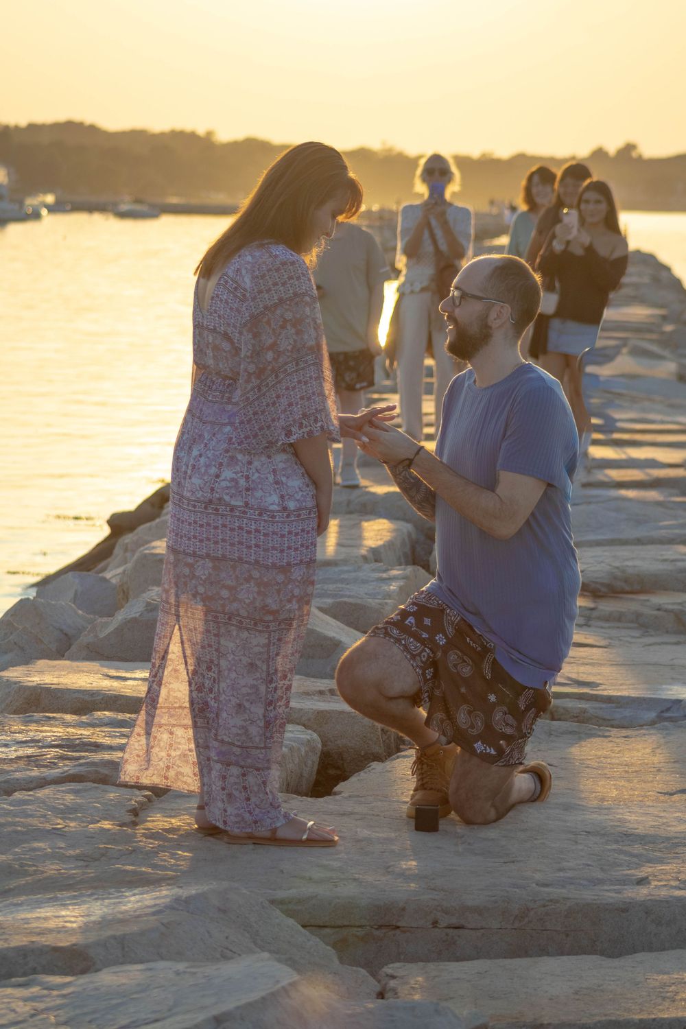 Man proposes to woman on rocky pier at sunset with onlookers nearby.