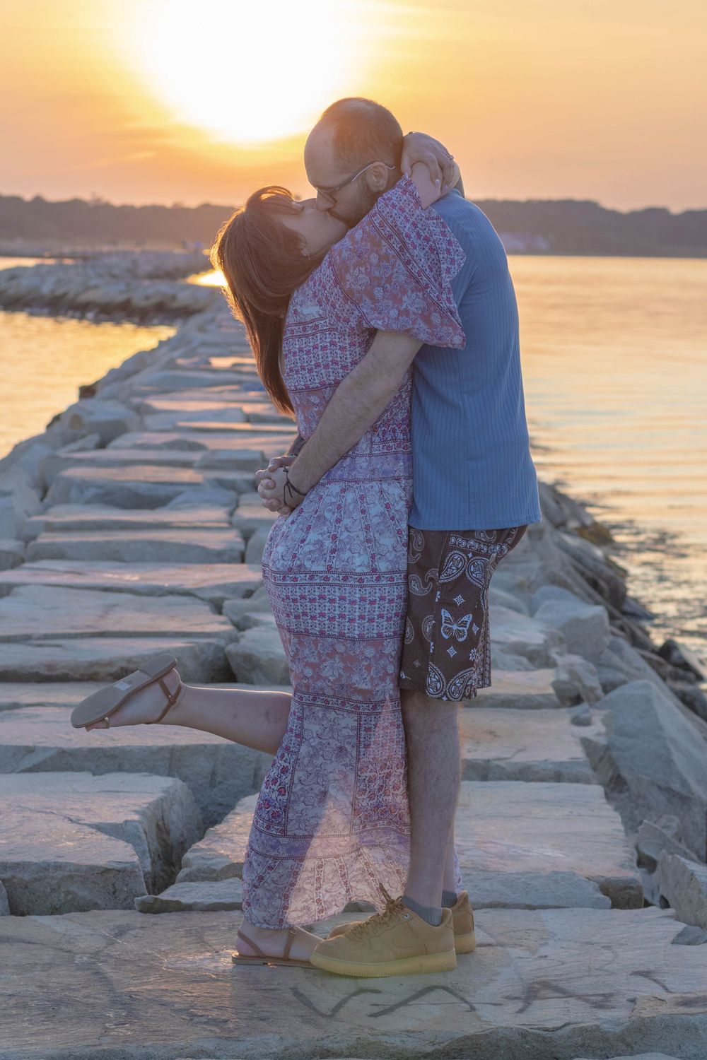 Couple sharing a romantic kiss on a rocky pier at golden sunset over calm water.