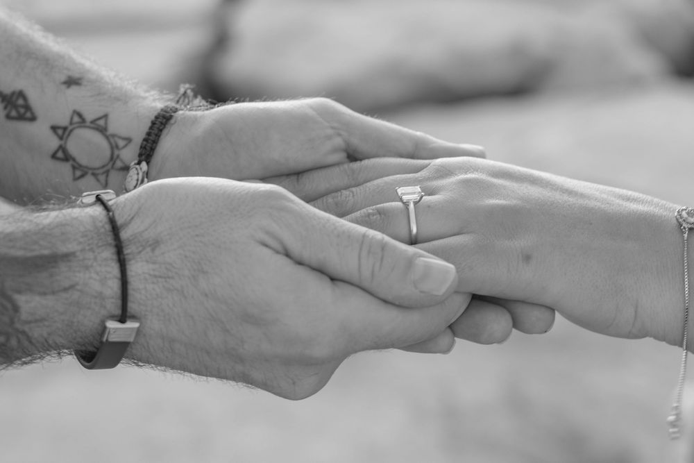 Black and white photo of two hands holding, one with a diamond engagement ring and the other with a tattoo and bracelet.