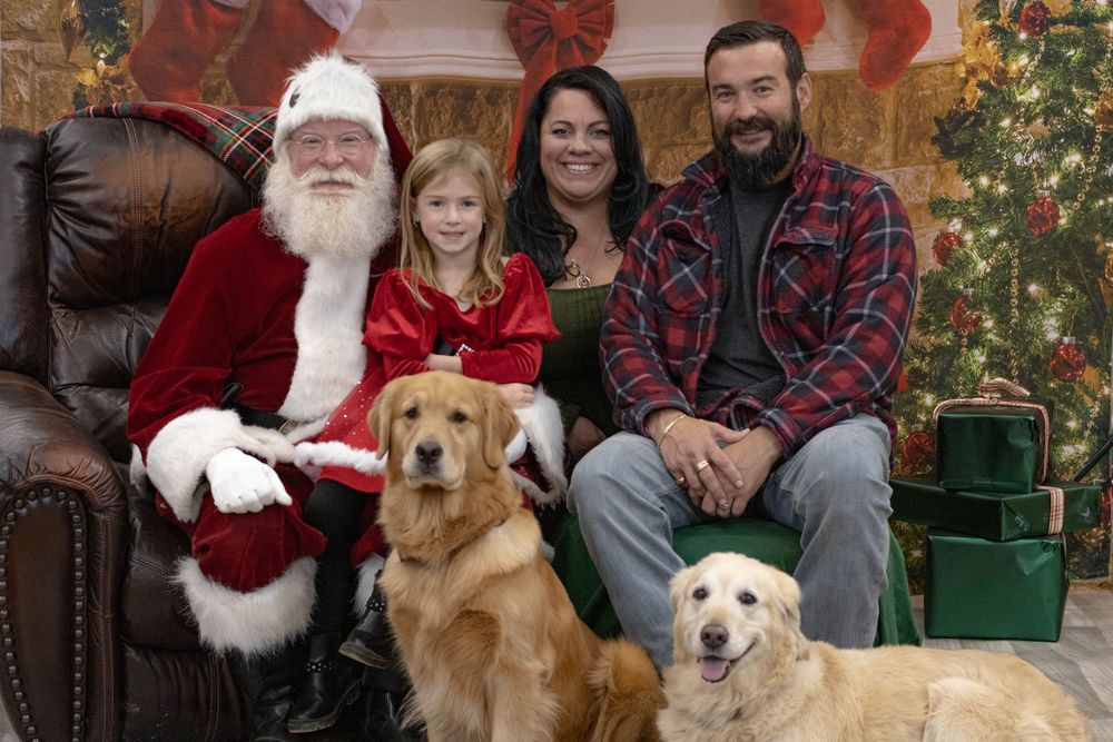 Family with two golden retrievers posing with Santa Claus in front of Christmas decorations and gifts.