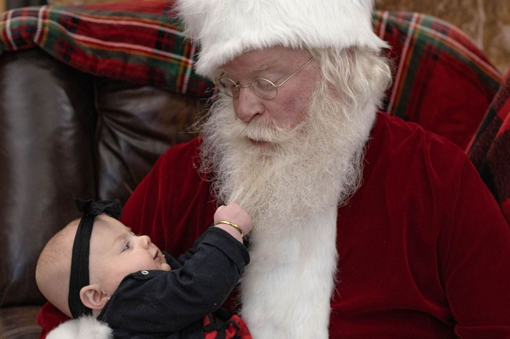 Newborn baby reaching up to touch Santa Claus beard during Christmas photo session.