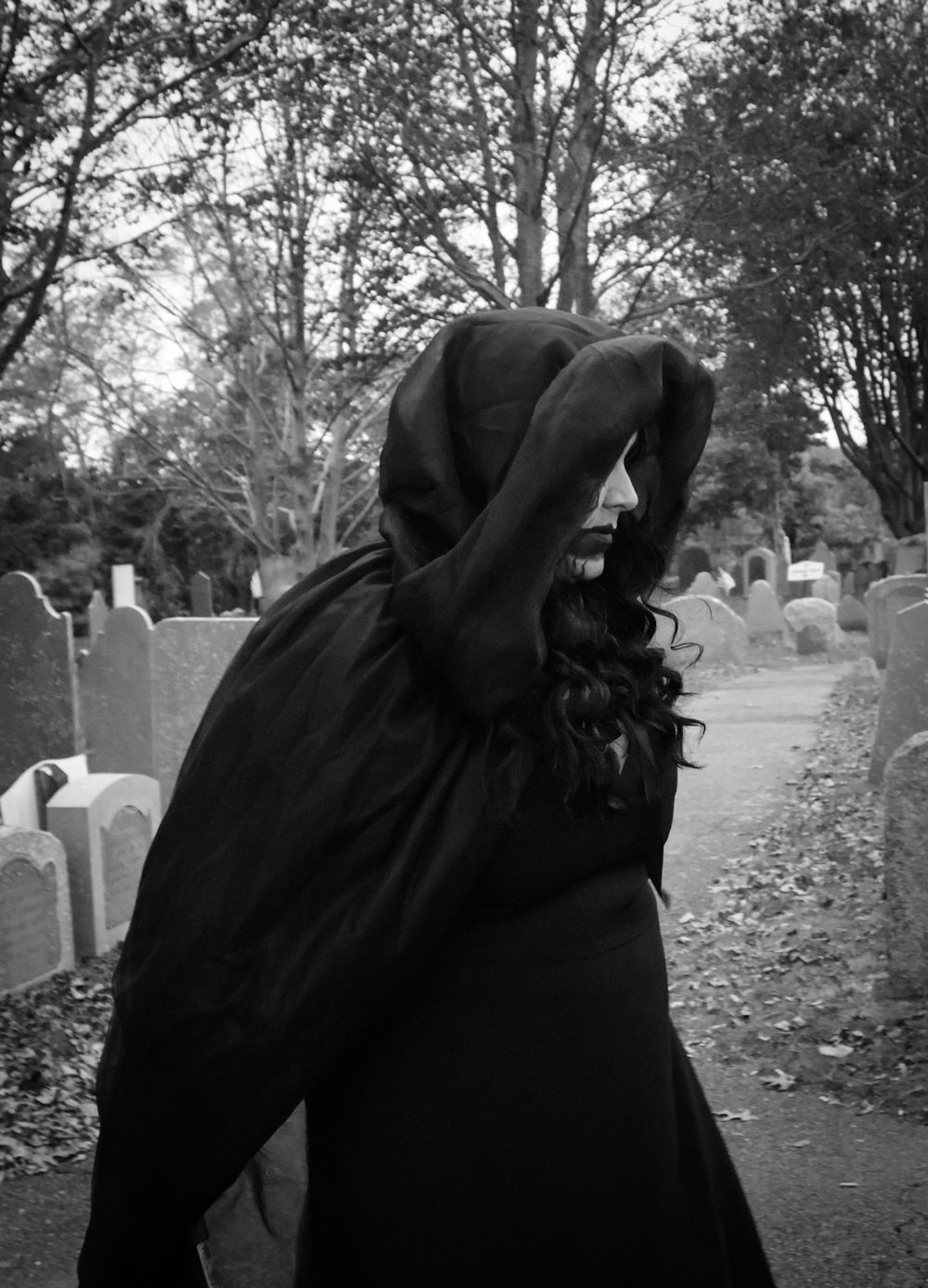Person in black hooded cloak standing in cemetery among gravestones, black and white gothic photography.