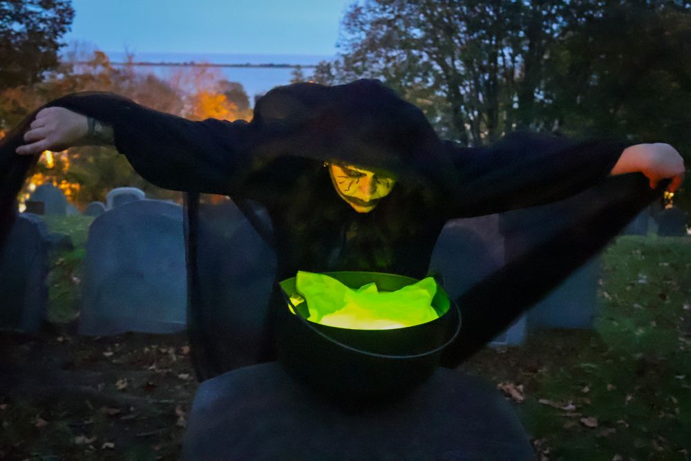 Person dressed as a witch in a cemetery at night, holding a glowing green cauldron among gravestones.