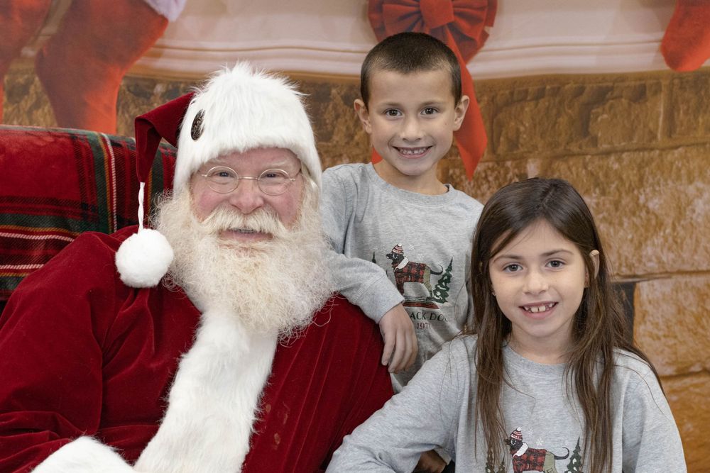 Two smiling children posing with Santa Claus in front of a festive Christmas fireplace backdrop.