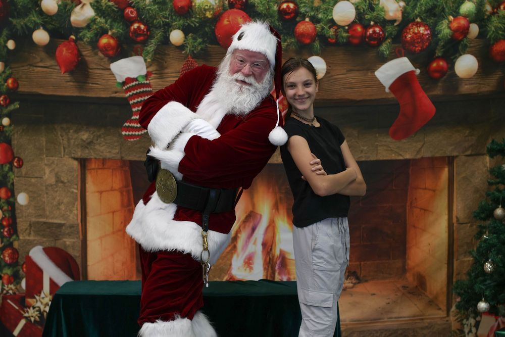 Young girl poses with Santa Claus in front of a decorated Christmas fireplace backdrop with stockings.