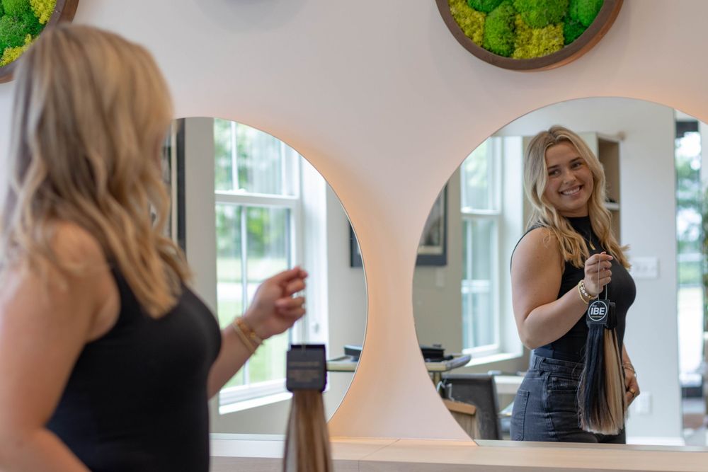 Blonde woman smiling in a hair salon mirror, holding hair extensions in a modern, stylish interior.