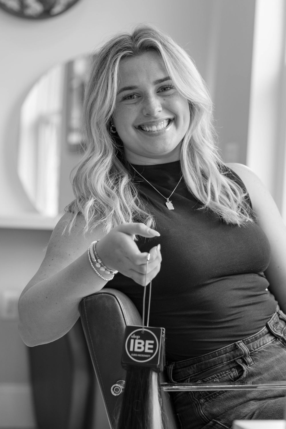 Smiling blonde woman holding an IBE hair extension tool in a black and white salon photo.