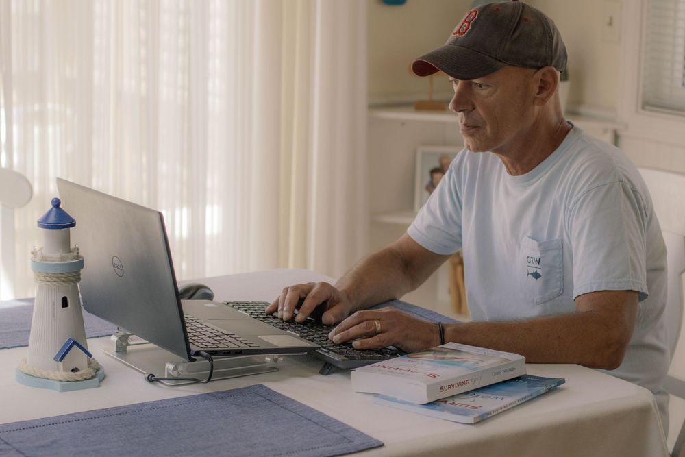 Senior man in baseball cap typing on laptop at home desk with decorative lighthouse figurine nearby.