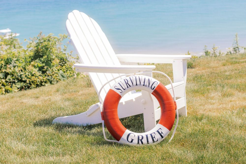 White Adirondack chair on green grass with a red and white life preserver ring reading Surviving Grief near blue water.