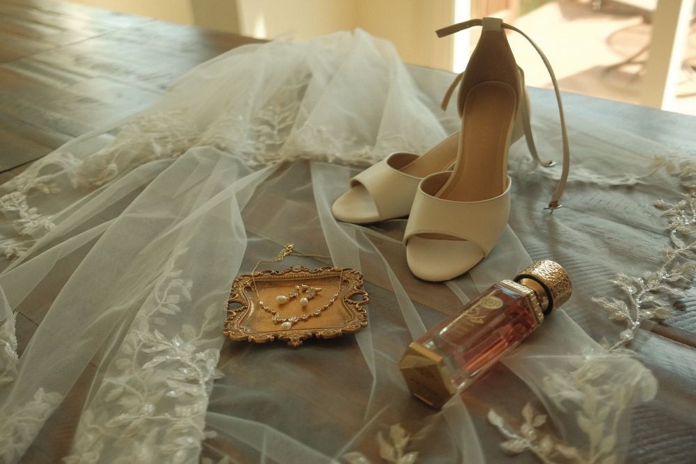 Bridal accessories flat lay featuring white heeled sandals, lace veil, gold jewelry box, and perfume bottle on a bed.