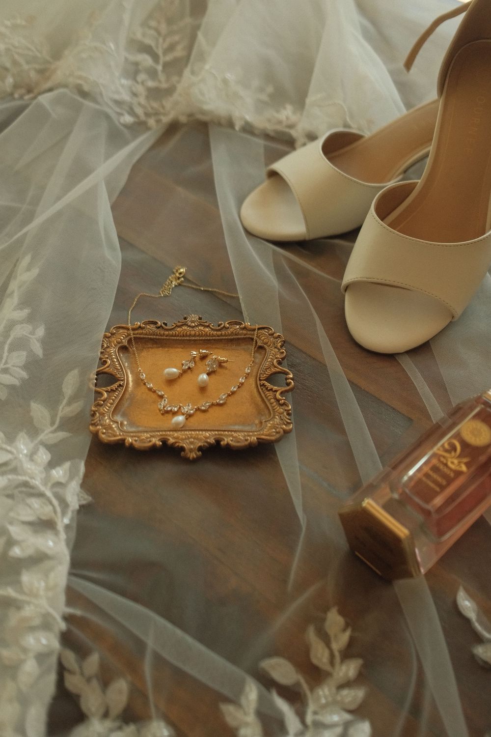 Elegant bridal flat lay with pearl jewelry in gold frame, nude heels, perfume bottle, and lace veil on wood surface.