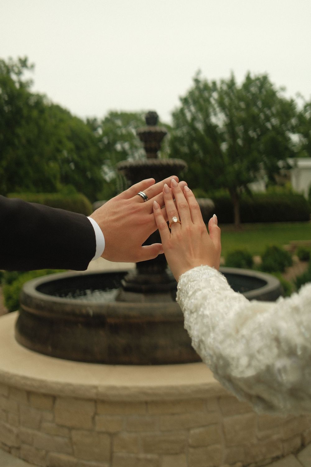 Newlyweds displaying wedding rings while touching hands near an elegant stone fountain in a garden.