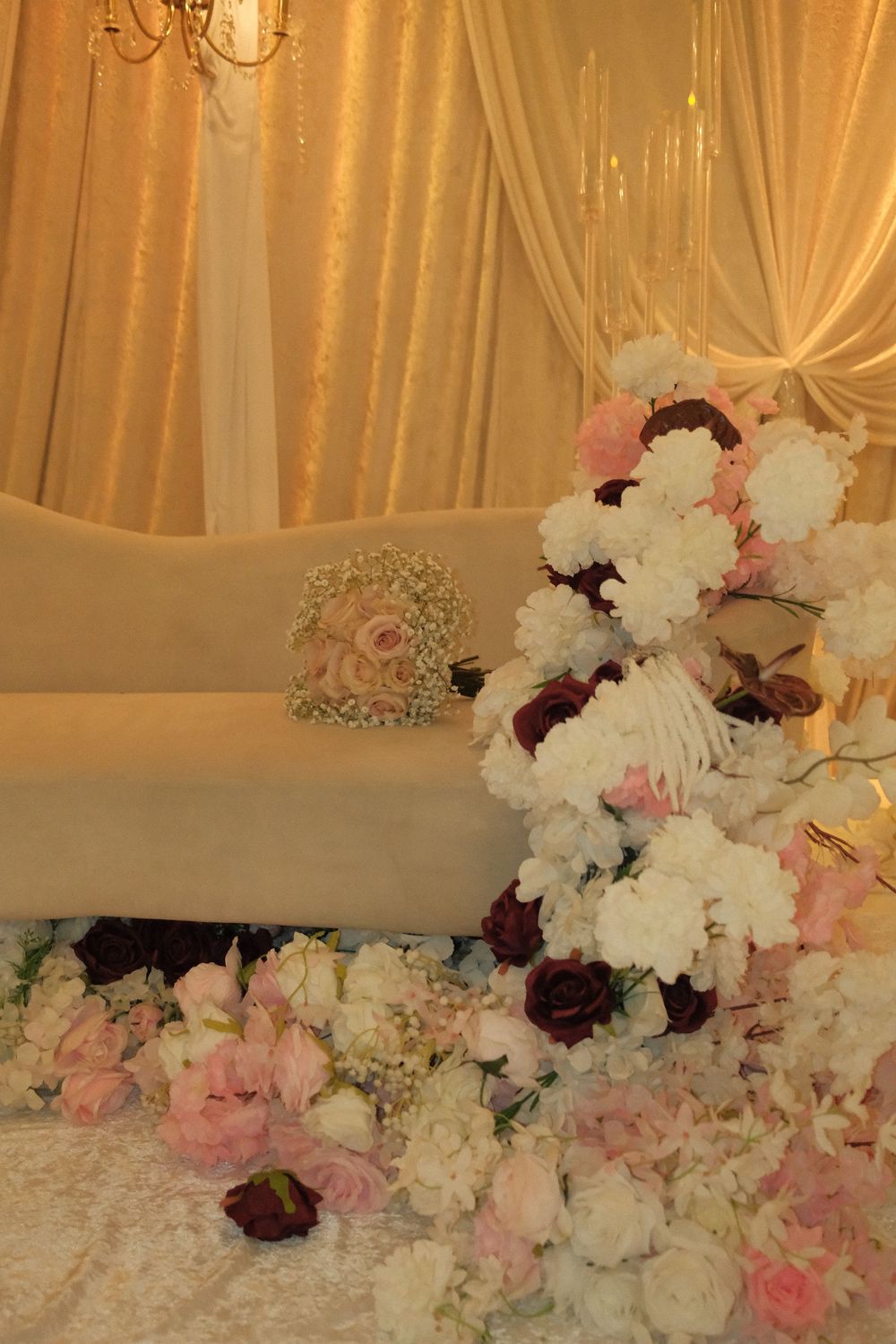 Elegant wedding reception setup with white and pink floral arrangements beside a decorated sofa under golden draping.