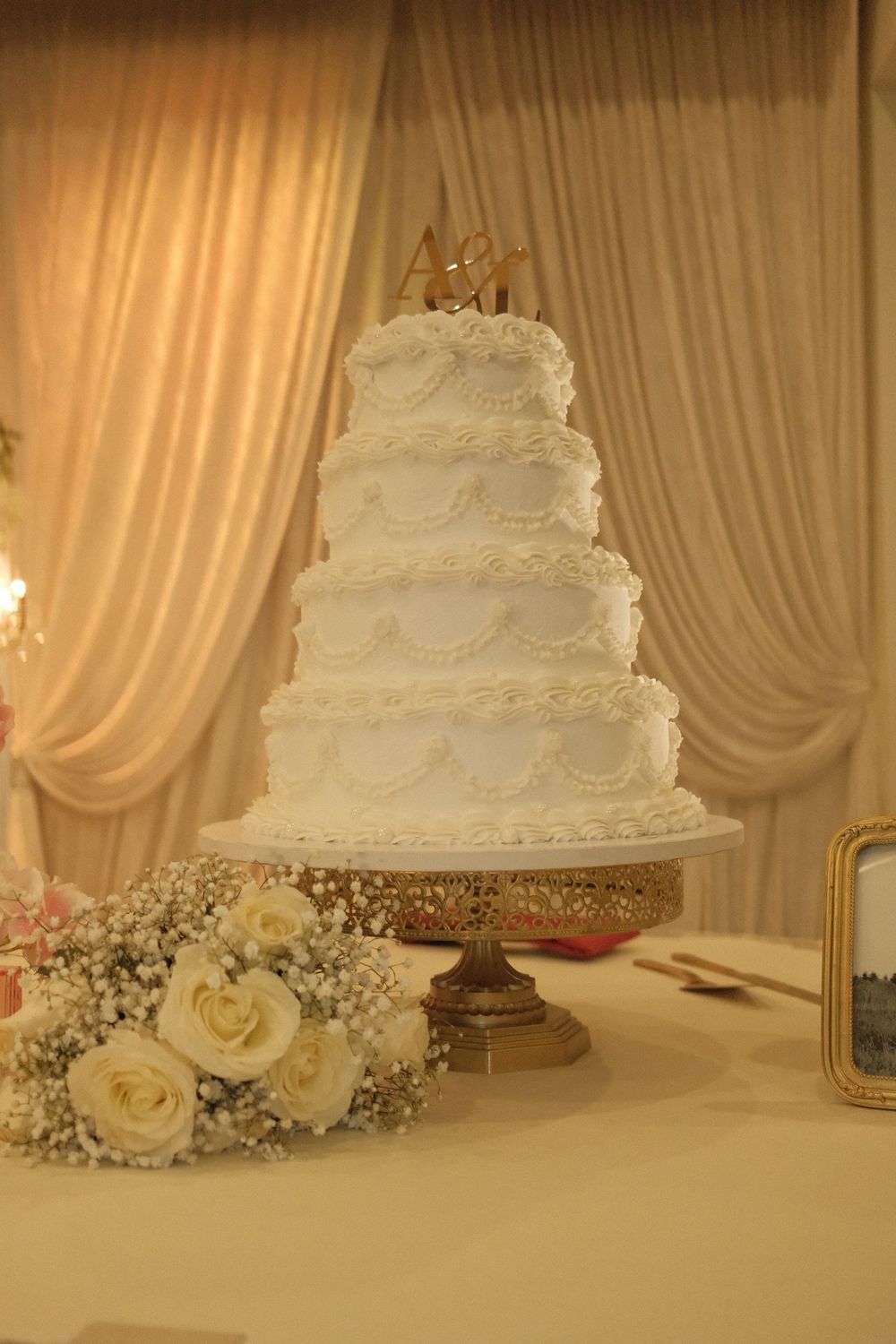 Elegant four-tier white wedding cake on gold stand with floral bouquet and draped curtain backdrop.
